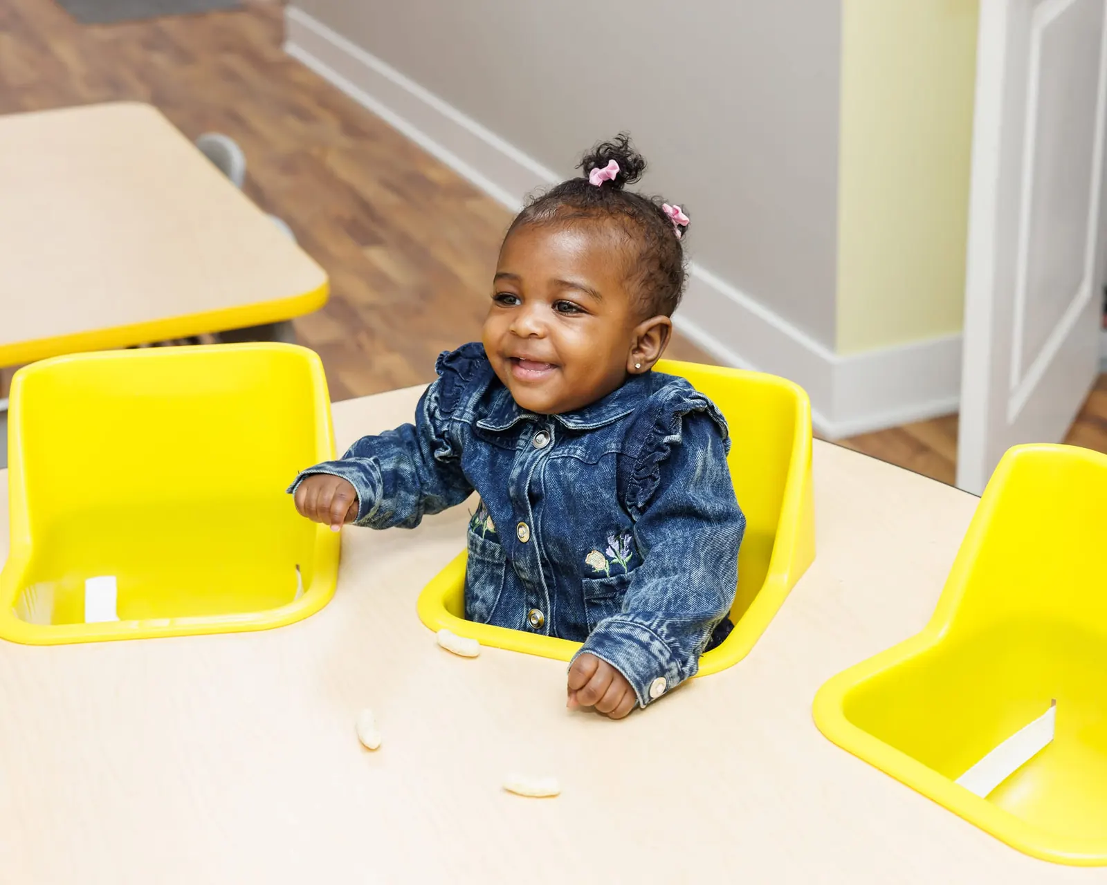 Smiling toddler in a denim jacket seated at a snack table with bright yellow chairs