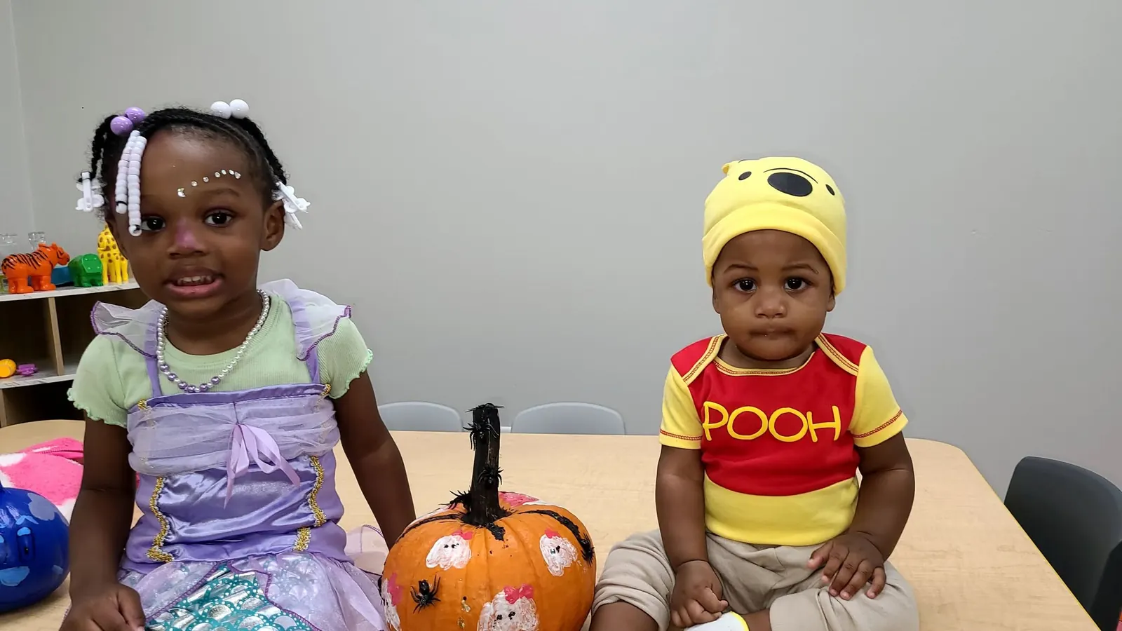 Two toddlers in costumes smiling beside a pumpkin